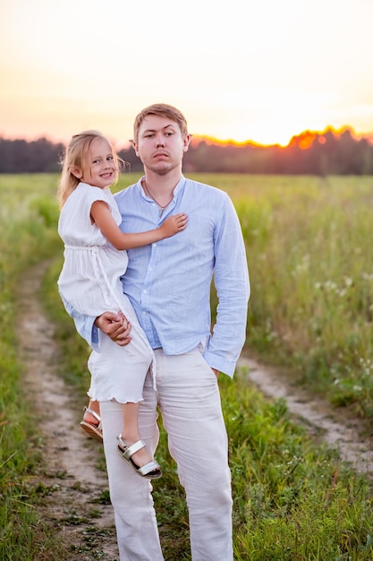 Little caucasian daughter in father's arms in the field on sunset summer time.