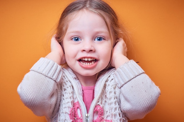 a little Caucasian girl covers her ears with her hands and smiles on a colored