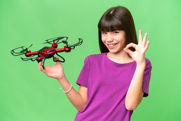 Little caucasian girl holding a drone over isolated background showing ok sign with fingers