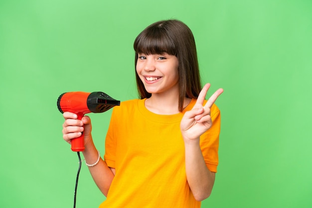 Little Caucasian girl holding a hairdryer over isolated background smiling and showing victory sign