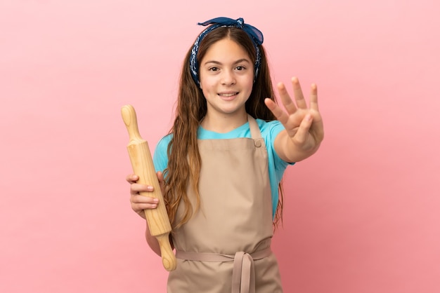 Little caucasian girl holding a rolling pin isolated on pink background happy and counting four with fingers