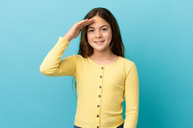 Little caucasian girl isolated on blue background saluting with hand with happy expression