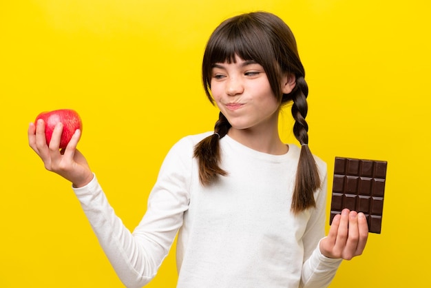 Little caucasian girl isolated on yellow background having doubts while taking a chocolate tablet in one hand and an apple in the other