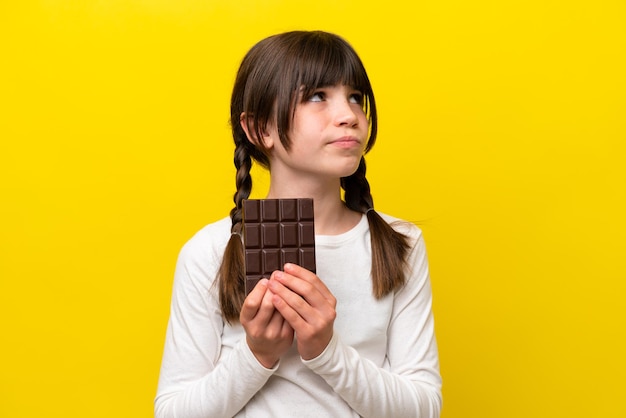 Little caucasian girl isolated on yellow background taking a chocolate tablet and having doubts