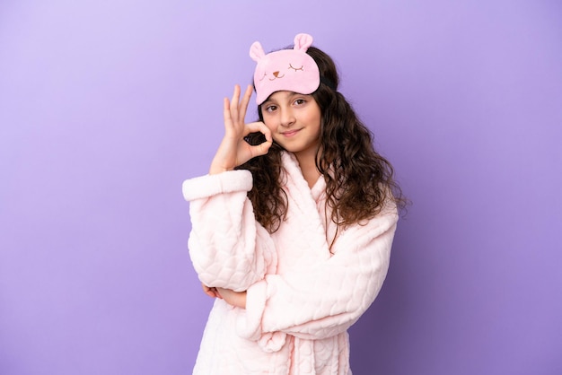 Little caucasian girl in pajamas isolated on purple background showing ok sign with fingers