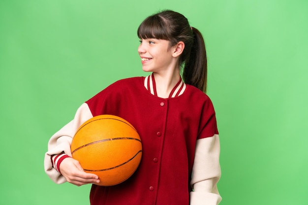 Little caucasian girl playing basketball over isolated background looking to the side and smiling