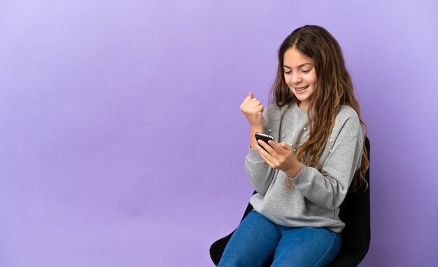 Little caucasian girl sitting on a chair isolated on purple background with phone in victory position