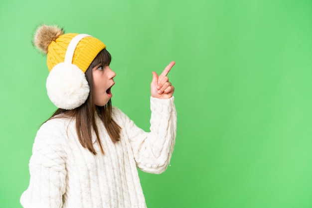 Little caucasian girl wearing winter muffs over isolated background pointing away