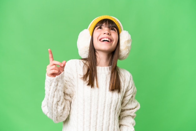 Little caucasian girl wearing winter muffs over isolated background pointing up and surprised