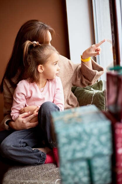 Little caucasian girl with mother looking at window and dreaming A lot of gift boxes around Happy family christmas concept