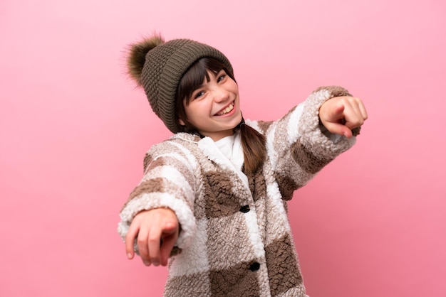 Little caucasian girl with winter jacket isolated on pink background points finger at you while smiling