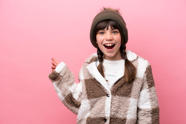 Little caucasian girl with winter jacket isolated on pink background surprised and pointing side