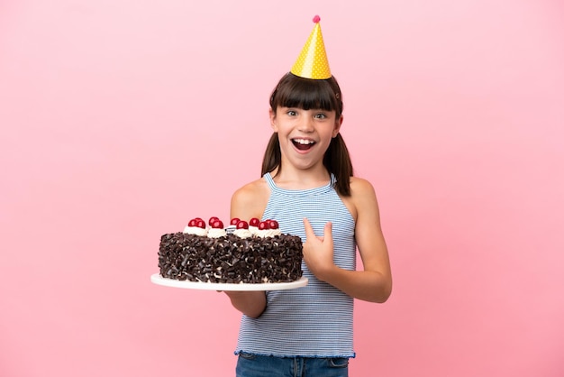 Little caucasian kid holding birthday cake isolated in pink background with surprise facial expression