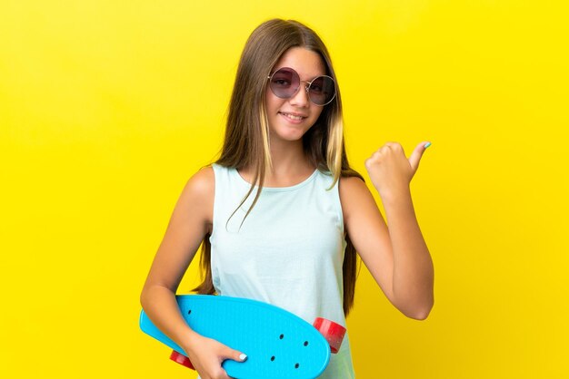 Little caucasian skater girl isolated on yellow background with a skate and pointing to the side