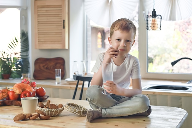 Little caucasian smiling boy sitting on the kitchen table and eat oatmeal cookies with milk