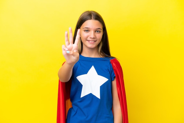 Little caucasian superhero girl isolated on yellow background happy and counting three with fingers