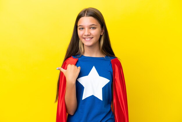 Little caucasian superhero girl isolated on yellow background pointing to the side to present a product