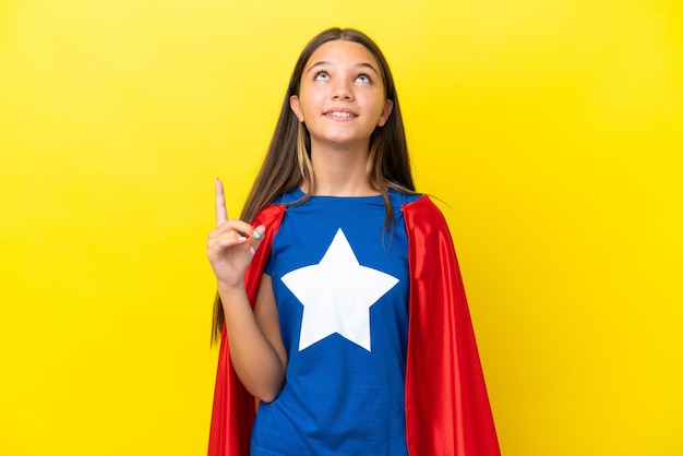 Little caucasian superhero girl isolated on yellow background pointing up and surprised