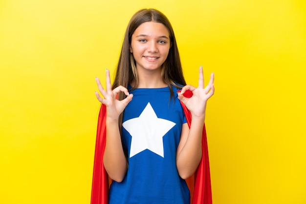 Little caucasian superhero girl isolated on yellow background showing an ok sign with fingers