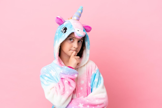 Little caucasian woman wearing a unicorn pajama isolated on pink background and looking up