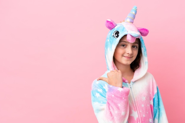 Little caucasian woman wearing a unicorn pajama isolated on pink background pointing to the side to present a product