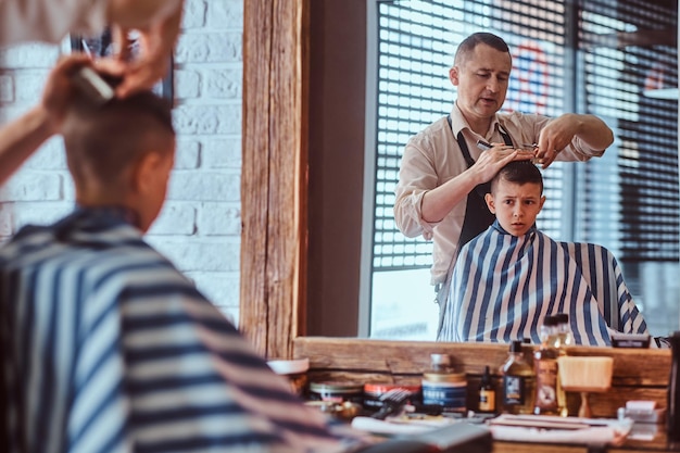 Little charming boy is getting trendy haircut from mature hairdresser at fashionable hairdressing salon.