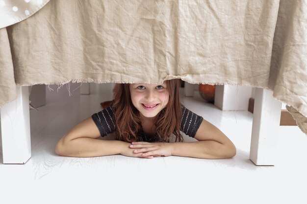 little cheerful girl climbs under the table and grimaces