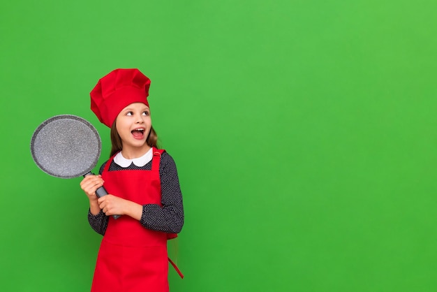 A little chef girl in a red hat and apron holds a pancake pan and looks at your advertisement on a green isolated background Choosing a profession for a student Copy space