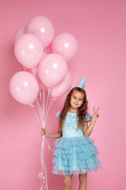 Little child girl in blue dress and birthday hat celebrating with pastel pink air balloons