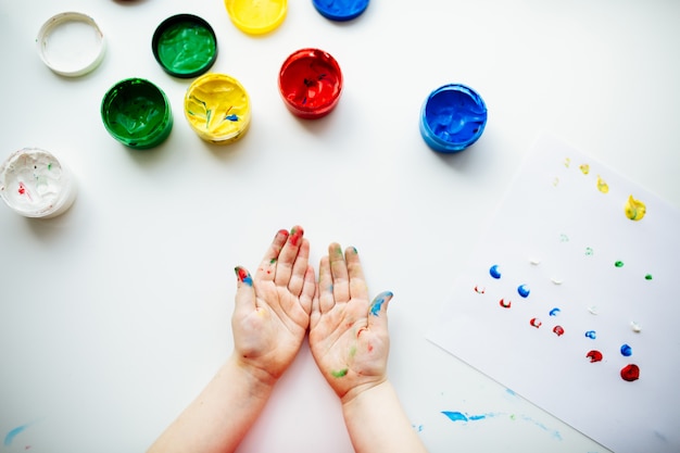 Little child shows his hands stained with paint at the table with art supplies, top view