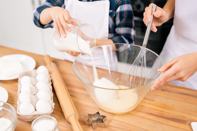 Little child sugar into bowl with shaked raw eggs while helping his mom with dough for homemade pastry by table in the kitchen