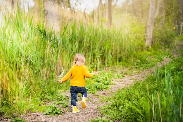 Little child wearing yellow rain boots, walking along a forest path in the grass 