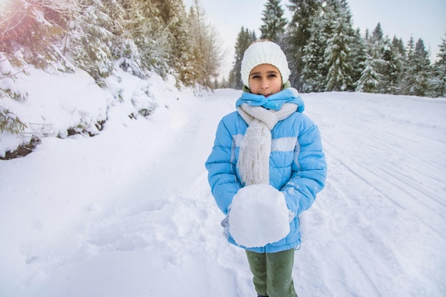 A little cunning cheerful girl in a sky blue jacket and a white knitted hat stands on a snowy road in a pine quiet winter forest and holds a large cold snowball