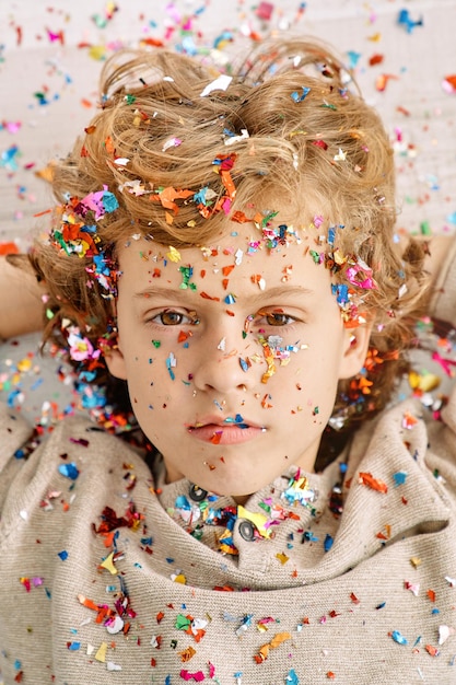 Little curly blond boy with colorful confetti looking at camera on white backdrop in studio