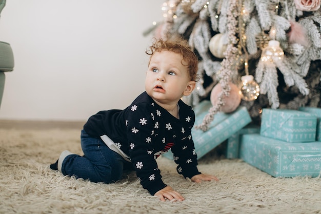 A little curly boy in a traditional Christmas sweater spends time at home near the Christmas tree