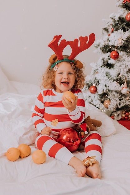 Little curly cheerful girl in christmas pajamas eats lollipop and tangerines sitting in bed child is