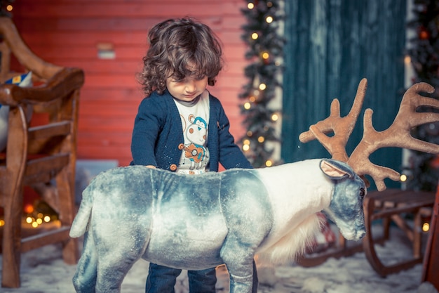 A little curly sweet kid boy in jeans playing with deer toy in the living room on Christmas. Family party