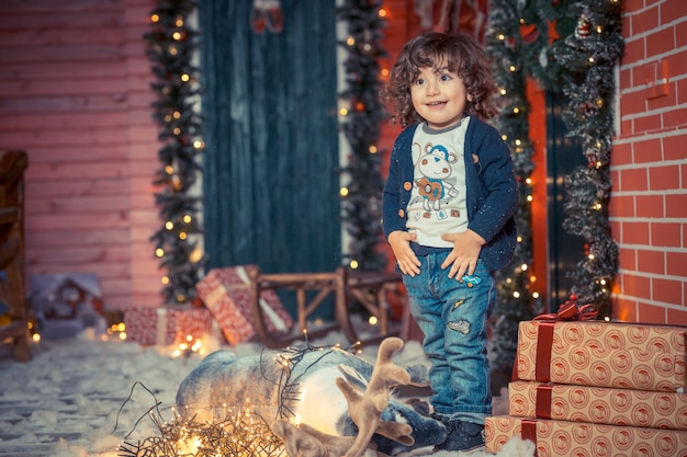 A little curly sweet kid boy in jeans standing near the deer toy and Christmas lights in the living room on Christmas