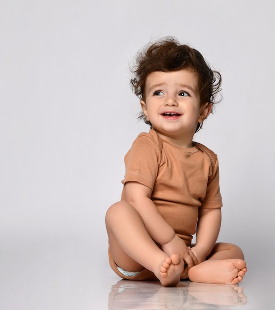 Little curlyhaired boy dressed in a brown bodysuit laughs out loud sitting barefoot on a gray background The notion of joy childhood and infancy