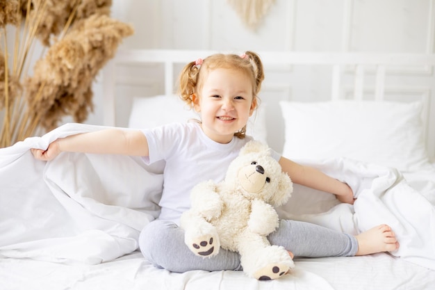 a little cute baby girl peeks out from under the blanket on a white cotton bed at home smiling hugging a teddy bear the child plays and indulges at home in the morning