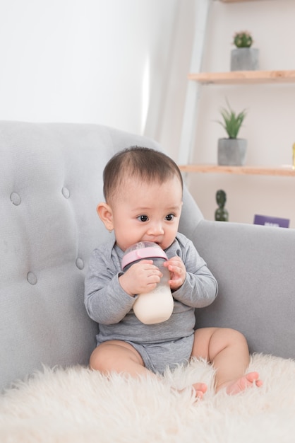 Little cute baby girl sitting in room on sofa drinking milk from bottle and smiling. Happy infant. Family people indoor Interior concepts. Childhood best time!