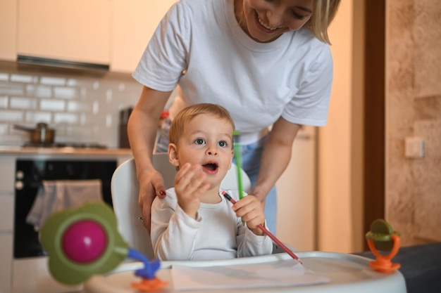 Little cute baby toddler boy blonde sitting on baby chair learning to draw beautiful young mom and