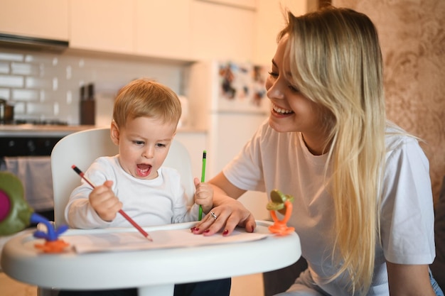 Little cute baby toddler boy blonde sitting on baby chair learning to draw beautiful young mom and