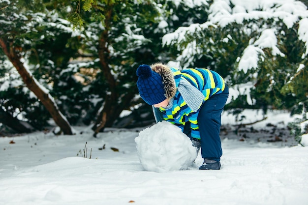 Little cute boy making snowman rolling big snowball