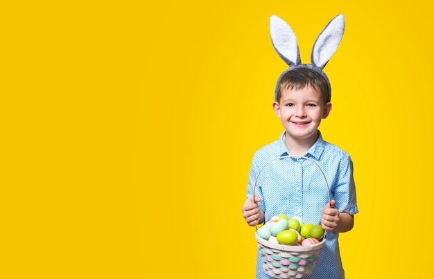 Little cute boy with an Easter basket in his hands and bunny ears on his head on a yellow background