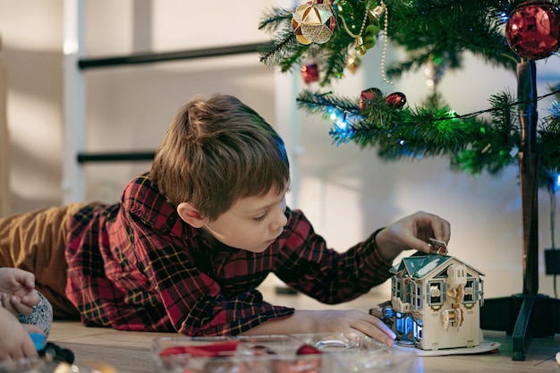 Little cute caucasian boy laying under Christmas tree at home playing with paper toy house