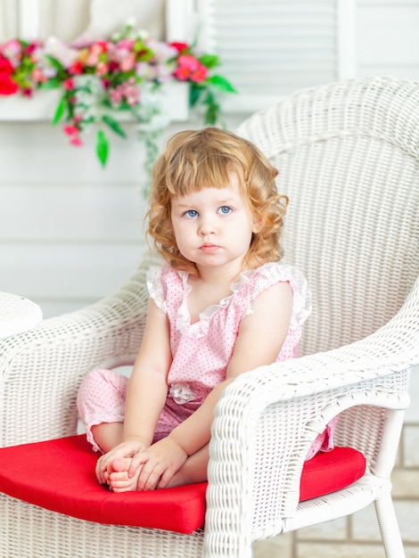 Little cute curly girl sitting on white wicker chair in the garden and looks into distance.