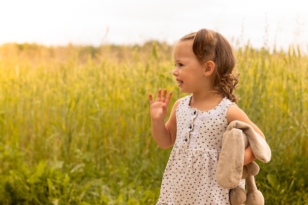 Little cute girl 1-3 with a plush hare in a light dress waving his hand in a field of spikelets of rye in summer