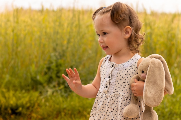 Little cute girl 1-3 with a plush hare in a light dress waving his hand in a field of spikelets of rye in summer