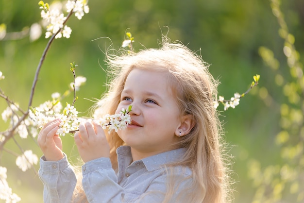 Little cute girl blonde with long hair sniffs a flowering tree branch in the park in spring.